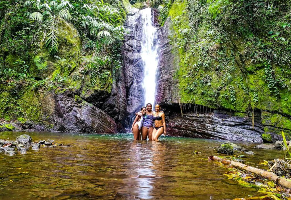 three black women friends pose standing in front of Bowden Hill waterfall with clear turquoise water in Jamaica